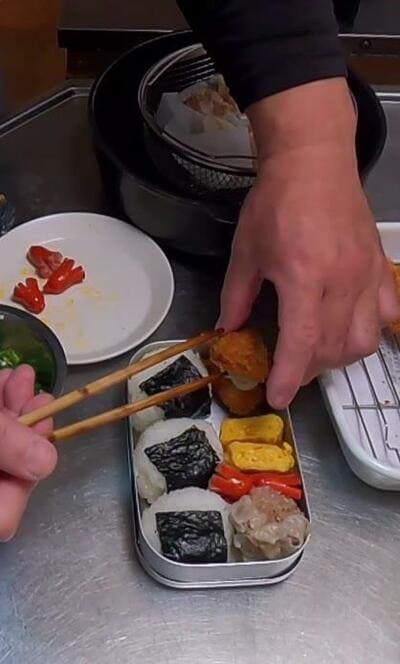 おにぎりやシウマイが並んだ弁当箱へ、太刀魚のフライを箸で詰める