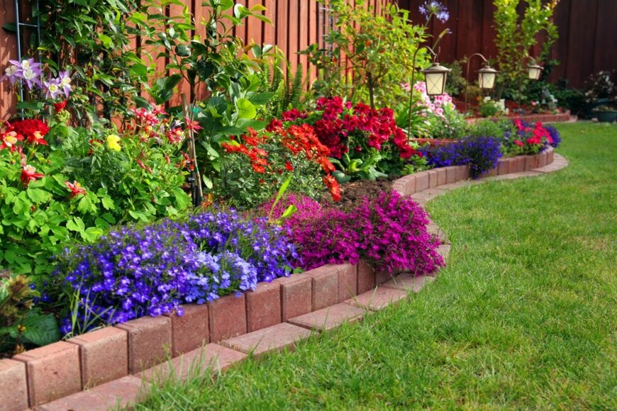 庭の花壇に花が植えられている。花の色は紫、赤、青など。花壇の仕切りはレンガ、手間には芝生が生えている。