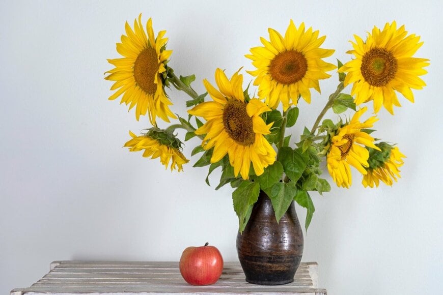 花瓶にヒマワリが活けられている。花瓶の左にはリンゴがある。