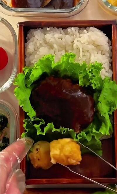 ハンバーグの手前に黄色く丸いおかずが置かれていくところ
